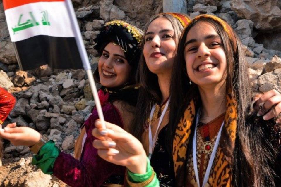 Young women dressed in traditional clothing wave an Iraqi national flag during Pope Francis 2021 visit to Iraq (AFP or licensors)